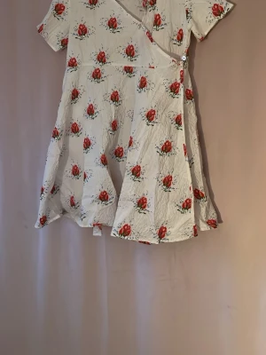 Vit kortklänning med röda rosor - Supersöt vit klänning med röda rosor och gröna blad i ett blommigt mönster. Klänningen har korta ärmar, omlott-detalj framtill och lätt utställd kjol. Perfekt för sommarens alla tillfällen och ger en romantisk vibe. storlek 7-8 år men passar 146/152 märke konges sløjd köpt på H10 på Gotland för 800kr (använd endast 2 gånger)