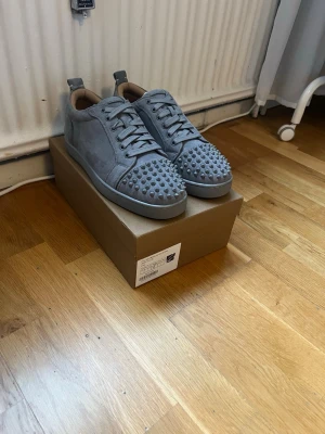 Gråa Christian Louboutin sneakers - Snygga gråa sneakers från Christian Louboutin i mocka med ikoniska nitar på tån och klassisk röd sula. Skorna har rundad tå, grå snörning och låg profil. Perfekta för dig som vill sticka ut med en lyxig och edgy stil.