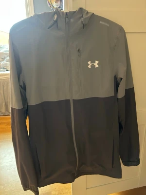 Ljus grå och mörk grå vindjacka Under Armour - Snygg tvåfärgad vindjacka från Under Armour i grått och svart. Jackan har huva, hel dragkedja framtill och en diskret logga på bröstet. Tillverkad i lätt och vindtätt material. Nypris 1200, oandvänd pga för liten 