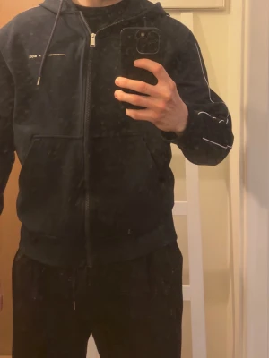 Svart hoodie Porsche x BOSS - Snygg svart hoodie från Porsche x BOSS- Limited Edtion, med dragkedja framtill och vita detaljer längs ärmarna. Hoodien har huva med dragsko och praktiska fickor. Perfekt för dig som gillar stilrena och exklusiva plagg. Köptes från emporia i Room 1006. Finns ej att köpa längre 