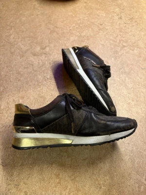 Bruna Michael Kors sneakers med gulddetaljer - Snygga svarta sneakers från Michael Kors med guldfärgade detaljer på häl och sula. Skorna har klassisk låg profil, snörning och en mix av läder och textilmaterial. Perfekta för dig som vill ha en stilren och trendig look med lite extra bling. Mycket sköna skor, men är slitna (syns på bilderna)