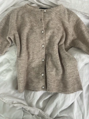 Beige kofta med pärlknappar - Mysig beige kofta med rund halsringning och pärlknappar framtill. Koftan har ribbade muddar vid ärmslut och nederkant samt en mjuk, stickad struktur. Perfekt för lager på lager och enkel att matcha med olika outfits.