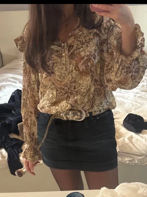 Mönstrad  guld blus med volangdetaljer - Säljer en bohemisk blus med guldiga och bruna mönster, volangdetaljer vid axlar och ärmslut samt knappar framtill. Blusen har långa ärmar och ett lätt glansigt material som ger en lyxig känsla. Perfekt för dig som gillar unika plagg med mycket detaljer.
