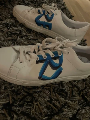 Burberry sneakers - Snygga vita sneakers från Burberry med en stor blå och svart logga på sidan. Skorna har klassisk låg profil, vita snören och sula i gummi. Perfekta för dig som vill ha en clean look med en twist.
