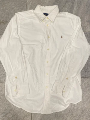 Vit skjorta från Ralph Lauren - Klassisk vit skjorta från Ralph Lauren med broderad logga på bröstet. Skjortan har lång ärm, knappar framtill och på manschetterna samt en snygg krage. Tillverkad i mjuk bomull som känns skön mot huden. Perfekt för en clean och stilren look.