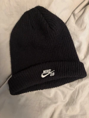 Svart ribbstickad mössa från Nike SB - Säljer en svart ribbstickad mössa från Nike SB med uppvikt kant och vit broderad logga framtill. Klassisk och enkel design som passar till det mesta. Perfekt för dig som gillar streetwear och vill hålla dig varm. Till både tjej och kille 