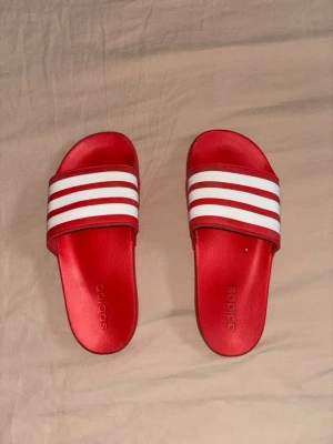 Röda Adidas badtofflor med vita ränder - Säljer ett par röda Adidas sandaler med klassiska vita ränder och Adidas-logga på remmen. Tillverkade i syntetmaterial med räfflad sula för bra grepp. Perfekta för stranden eller poolen och har en sportig look. Är dock lite smutsiga men går antagligen att skölja bort.