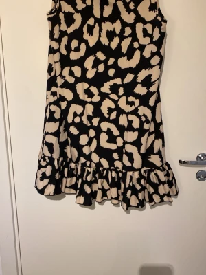Leopardmönstrad ärmlös kortklänning - Säljer en ärmlös kortklänning med beige och svart leopardmönster. Klänningen har volangkant nedtill och en dekorativ rosett i nacken. Perfekt för dig som vill sticka ut med ett djurmönster och gillar en loose passform.