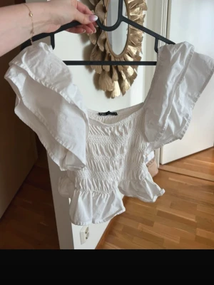 Vit topp med volangärmar - Säljer en söt vit topp med breda volangärmar och smockad midja. Toppen har en kort, figurnära passform och rund halsringning. Perfekt för dig som gillar romantisk och trendig stil.