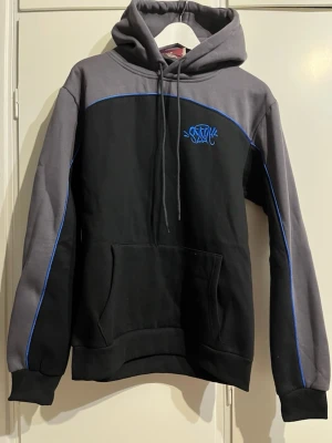 Svart och grå hoodie med blå detaljer - Snygg hoodie i svart och grått med blåa sömmar och broderad logga på bröstet. Huva med dragsko och stor magficka framtill. Perfekt för dig som gillar streetwear och vill ha en stilren men ändå unik look.