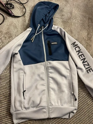 Blå och grå hoodie från McKenzie - Snygg hoodie från McKenzie i blått och ljusgrått med dragkedja framtill, huva med snörning och fickor med dragkedja. Stor McKenzie-logga på ena ärmen och bröstficka med dragkedja. Perfekt för en chill och sportig stil.