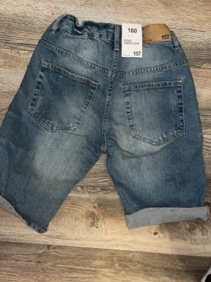Blå denimshorts med snörning - Snygga blå denimshorts från 157 med snörning i midjan och uppvikta benslut. Shortsen har fem fickor, slitningar framtill och en avslappnad passform. Perfekta för varma dagar och enkel att matcha med olika stilar.
