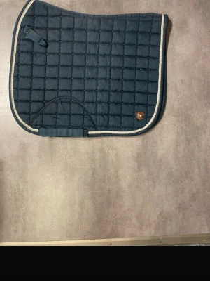 Mörkblått schabrak med kantband - Säljer ett mörkblått schabrak från Trinty med rutmönster och ljusbeige kantband. Schabraket har en liten logga i hörnet och praktiska fästen för sadel och gjord. Perfekt till ridning och ger en snygg look till din häst.