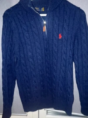 Mörkblå kabelstickad tröja Polo Ralph Lauren - Kabelstickad mörkblå tröja från Polo Ralph Lauren med dragkedja och hög krage. Tröjan har det klassiska röda Polo-logot på bröstet och ribbade muddar. Perfekt för dig som gillar stilren och tidlös design. Den är i bra skick men är lite nopprig. Storleken är L men den sitter som M