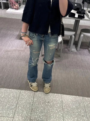 Blå slitna bootcut jeans - Säljer ett par blå bootcut jeans med slitningar och hål på knäna. Jeansen har en klassisk femficksmodell och är tillverkade i denim. Perfekta för en avslappnad och trendig look. Köp snabbt så kan priset diskuteras.
