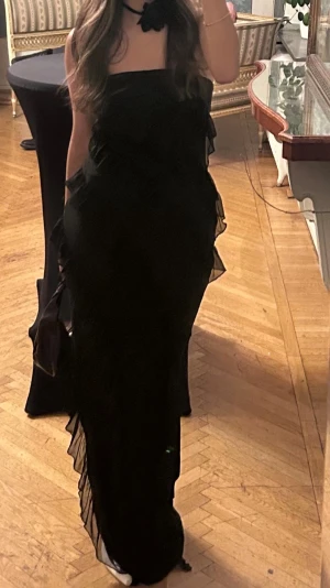 Svart långklänning med volanger - Säljer en svart axelbandslös långklänning med volangdetaljer i flera lager. Klänningen har en elegant och figurnära passform med asymmetriska volanger som ger en cool och trendig look. Perfekt för dig som vill sticka ut på nästa event.