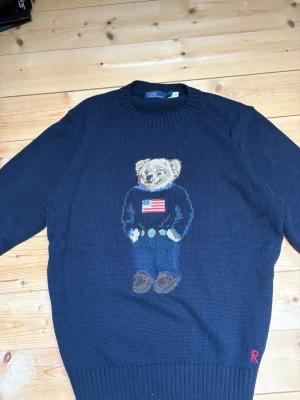 Mörkblå stickad tröja Polo Bear - Mörkblå stickad tröja från Polo Ralph Lauren med den ikoniska Polo Bear på bröstet, klädd i jeans och tröja med amerikanska flaggan. Rund halsringning och ribbade muddar. RL-broderi i rött nedtill. Perfekt för dig som gillar klassisk preppy stil.