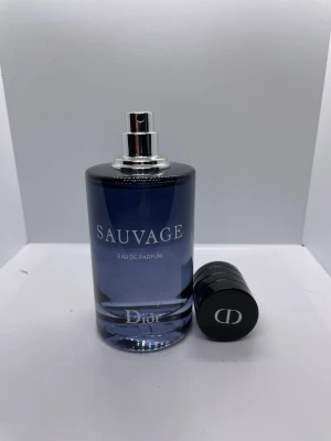 Dior Sauvage Eau de Parfum - Dior Sauvage Eau de Parfum med en elegant mörkblå glasflaska och silverdetaljer. Flaskan har rund form och svart lock med CD-logga. Kommer i en stilren svart kartong med vit text. En ikonisk doft för dig som vill sticka ut.