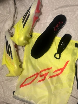 Adidas F50 Elite LL AG fotbollsskor - Säljer ett par Adidas F50 Elite LL AG fotbollsskor i gul och svart med röda detaljer. Skorna har låg vikt, strömlinjeformad design och är tillverkade i syntetmaterial. Perfekta för spel på konstgräs. Medföljer innersula, skohorn och F50-påse.