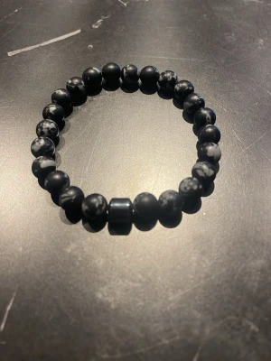 Svart pärlarmband med stenmönster - Snyggt armband med svarta pärlor och gråspräckliga detaljer. Pärlorna är runda och sitter på en elastisk tråd, vilket gör det enkelt att ta på och av. Perfekt accessoar för dig som gillar stilrena och unika smycken.