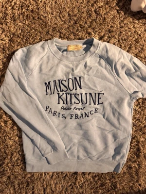 Ljusblå Maison Kitsuné sweatshirt 🙌🤩 - Ljusblå sweatshirt från Maison Kitsuné med broderad text 'Palais Royal Paris, France' på bröstet. Klassisk passform med rund hals och ribbade muddar. Perfekt för en chill och stilren look.🙌🤩