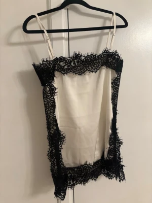 Vit sidenlinnetopp med svart spets - Snygg vit linnetopp i siden med tunna axelband och bred svart spets runt kanterna. Toppen har en elegant och lyxig känsla med sitt glansiga material och kontrasterande spetsdetaljer. Perfekt för att skapa en chic look.