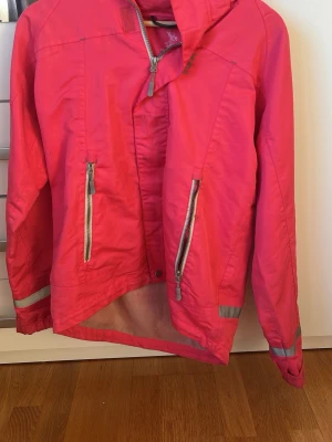 Rosa regnjacka med reflexdetaljer - Snygg rosa regnjacka med huva och två dragkedjeförsedda fickor framtill. Jackan har reflexdetaljer på ärmar och rygg för extra synlighet. Tillverkad i ett vattenavvisande material och har meshfoder på insidan för bättre komfort.
