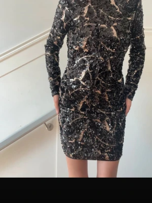 Svart glittrig paljettklänning - Snygg svart kortklänning täckt i paljetter med lång ärm och öppen rygg med korsade band. Klänningen har en figurnära passform och ett glittrigt mönster i silver och beige toner. Perfekt för dig som vill sticka ut.
