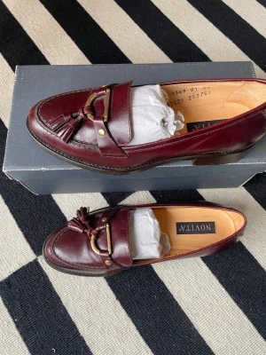 Vinröda ballerinaskor med tofsar från Novita - Snygga vinröda loafers från Novita i äkta läder. Skorna har dekorativa tofsar och guldfärgade detaljer framtill. Klassisk design med låg klack och rundad tå. Perfekta för dig som vill ha en stilren och tidlös look. 