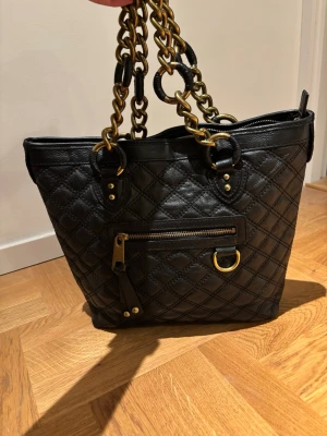 Svart quiltad Marc Jacobs tote stambag  med kedjor - Snygg svart handväska i quiltat läder med guldfärgade kedjehandtag. Väskan har en dragkedjeficka framtill och dekorativa metalldetaljer. Klassisk form med rymligt innerfack och coola detaljer som ger en lyxig känsla.