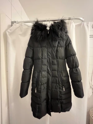 Svart vinterjacka från Roco Baroco storlek 34 - Svart lång jacka från Roco Baroco med snygg pälsdetalj på huvan. Jackan har quiltad design, dragkedja framtill och två fickor med dragkedja. Dragkedjan längst ner har lossnat i sömmarna men kan fixas lätt, därav priset. Nypris 2499kr. 