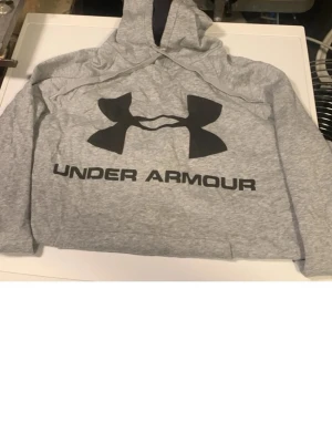 Grå hoodie från Under Armour - Snygg grå hoodie från Under Armour med stor svart logga och text på bröstet. Klassisk modell med huva och magficka. Perfekt för en sportig och avslappnad stil. Mjuk och bekväm bomullsblandning. Står storlek LG men skulle säga att den är mellan medium och large 