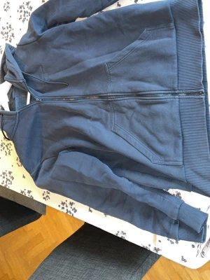 Blå hoodie från Peak Performance - Snygg blå hoodie från Peak Performance med dragkedja framtill och två stora fickor. Broderad logga på bröstet och klassisk huva med dragsko. Tillverkad i mjukt bomullsmaterial och har ribbade muddar vid ärmslut och nederkant. Perfekt för en chill och sportig look.