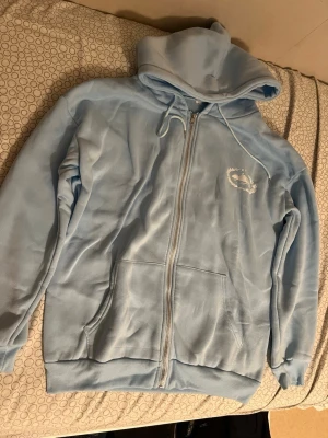 Ljusblå hoodie med dragkedja - Ljusblå hoodie med dragkedja och fickor framtill. Har en vit broderad logga på bröstet och justerbar huva med snören. Tillverkad i mjukt material som känns skönt mot huden. Perfekt för en avslappnad och trendig look. Corteiz