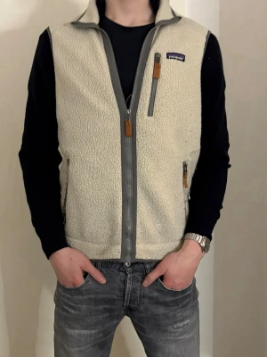Beige fleeceväst från Patagonia - Snygg beige fleeceväst från Patagonia med hel dragkedja framtill och tre fickor med dragkedja. Västen har en mjuk, teddy-liknande yta och detaljer i grått samt bruna dragkedjehängen. Perfekt för lager-på-lager och chill stil.