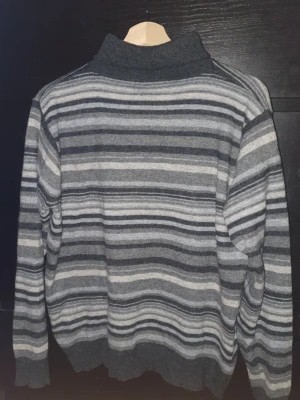 Gråmelerad stickad tröja med ränder - Säljer en stickad tröja i olika nyanser av grått med horisontella ränder. Tröjan har hög krage och ribbstickade muddar vid ärmslut och nederkant. Inte super tjock så lätt att layer och ha i garderoben. 