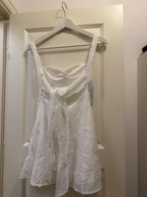 Vit Mcbeath kort klänning - Säljer en vit kort klänning från Mcbeath med broderade blommor och smala axelband. Var menad att ha som student klänning, dock köpt i för liten storlek US 0, alltså XS. Helt oanvänd med lappar kvar och i originalpaket.