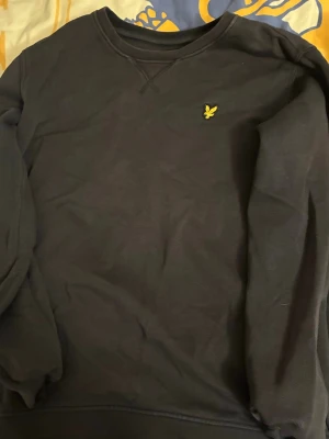 Svart sweatshirt från Lyle & Scott - Cool svart sweatshirt från Lyle & Scott med klassisk rund hals och gul logotyp på bröstet. Tröjan har ribbade muddar vid ärmslut och nederkant, perfekt för en avslappnad look. Materialet är mjukt och bekvämt. Nypris ca 599kr