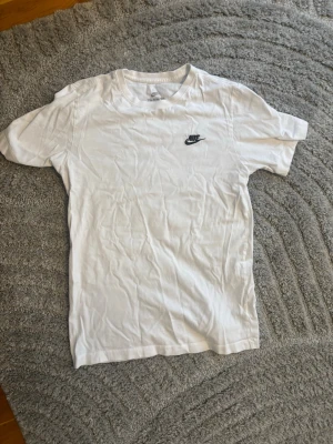 Vit Nike t-shirt - Klassisk vit t-shirt från Nike med liten svart logga broderad på bröstet. T-shirten har rund halsringning och korta ärmar. Perfekt basic-plagg för en avslappnad stil.