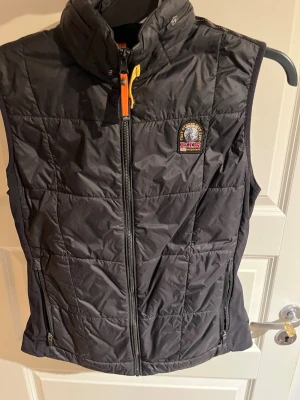 Svart dunväst från Parajumpers - Snygg svart dunväst från Parajumpers med hög krage, dragkedja framtill och två fickor med dragkedja. Västen har ett broderat Parajumpers-märke på bröstet och gul detalj vid dragkedjan. Perfekt för lager-på-lager och streetwear. Finns även en tunn luva som går att fälla in och ut med hjälp av dragkedja.