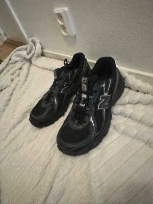 Svarta New Balance sneakers - Säljer ett par svarta New Balance sneakers med silvriga detaljer och meshpaneler för extra ventilation. Skorna har en chunky sula och klassisk snörning. Perfekta för dig som gillar sportig och stilren look.