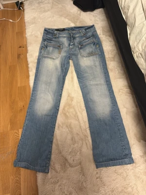 Ljusblå bootcut jeans med slitningar - Säljer ett par ljusblå bootcut jeans med slitningar och snyggt tvättade detaljer. Jeansen har fem fickor, dragkedja och knapp framtill samt dekorativa sömmar. Materialet är klassisk denim i bomull. Perfekta för en avslappnad och trendig look.