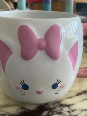 Söt kattmugg med rosa rosett - Unik mugg i keramik med kattmotiv, stora blå ögon och rosa detaljer som öron, nos och en stor rosett. Perfekt för te eller kaffe och har ett gulligt ansikte på framsidan. Muggen är vit med rosa handtag och passar dig som gillar söta och lekfulla saker.