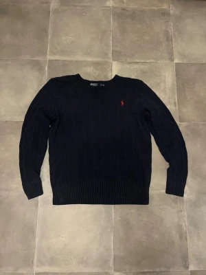 Mörkblå stickad tröja från Polo Ralph Lauren - Klassisk mörkblå stickad tröja från Polo Ralph Lauren med rund halsringning och diskret röd logga broderad på bröstet. Tröjan har kabelstickat mönster, ribbade muddar vid ärmslut och nederkant samt en avslappnad passform.