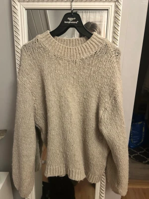 Beige stickad tröja med grov struktur - Mysig beige stickad tröja från Bagheera med grovstickad struktur och bred ribbad halsringning. Tröjan har avslappnad passform, långa ärmar och ribbade muddar vid ärmslut och nederkant. Perfekt för lager på lager under kyliga dagar.
