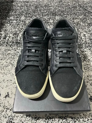  Saint Laurent sneakers - Svarta låga sneakers från Saint Laurent med vit logga på sidan. Ovandelen är i canvas och läder, med svarta skosnören och vit sula. Klassisk och stilren design som passar till det mesta. Perfekta för dig som gillar exklusiva streetwear-vibbar.