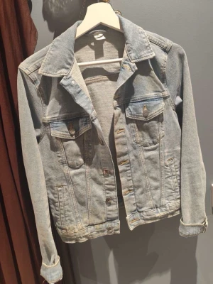 Ljusblå klassisk jeansjacka - Snygg ljusblå jeansjacka med klassisk krage, två bröstfickor med lock och knapp samt raka sömmar. Jackan har knappstängning framtill och justerbara ärmslut. Perfekt att slänga över en hoodie eller t-shirt för en avslappnad look.