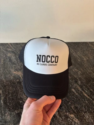 Svartvit NOCCO keps - Snygg svartvit truckerkeps från NOCCO med tydlig logga framtill. Kepsen har vit frontpanel, svart skärm och svart mesh baktill för extra ventilation. Justerbar baktill för perfekt passform. Perfekt för dig som gillar sportig och avslappnad stil. Helt ny aldrig använt den