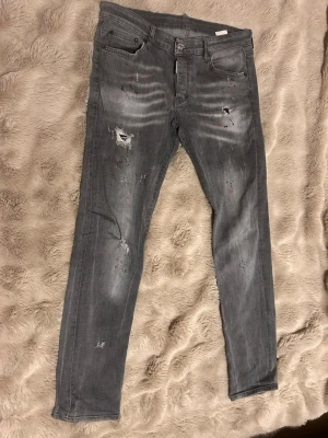 Grå slitna jeans från Dsquared2 - Säljer ett par grå jeans från Dsquared2 med skinny fit och slitningar, hål och färgstänk för en riktigt trendig look. Jeansen har fem fickor, knappgylf och klassisk läderpatch bak i midjan. Perfekta för dig som gillar streetstyle och edgy detaljer.