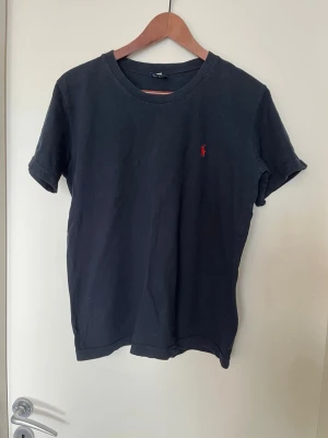 Svart t-shirt från Ralph Lauren - Klassisk svart t-shirt från Ralph Lauren med rund halsringning och korta ärmar. Liten röd broderad logga på bröstet. Tillverkad i mjuk bomull för en skön känsla och enkel stil.